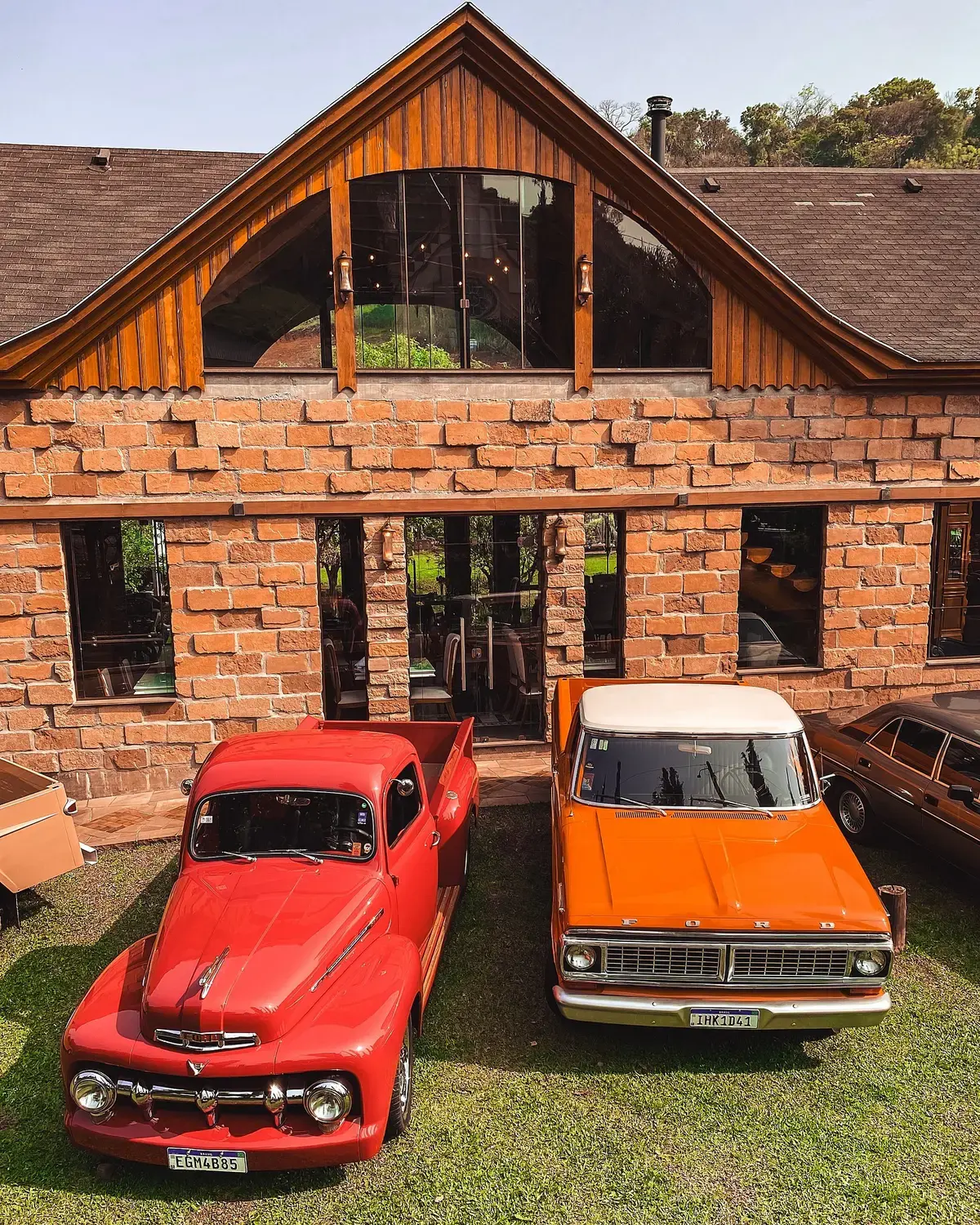 Duas picapes Ford vermelha e laranja estacionadas diante de uma construção de tijolos.
