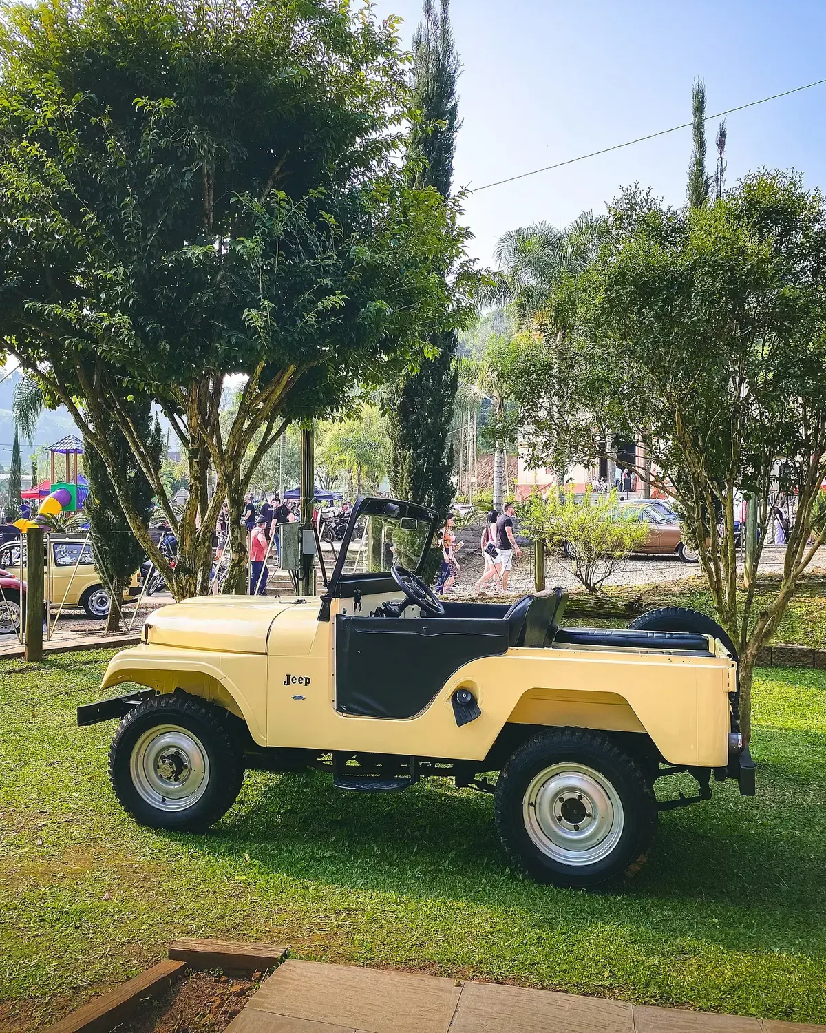 Jeep amarelo sem capota estacionado sob árvores durante um encontro de carros antigos.