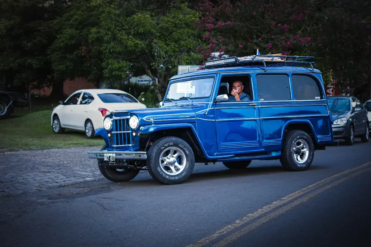 Rural Willys azul em movimento por uma rua arborizada durante um encontro.