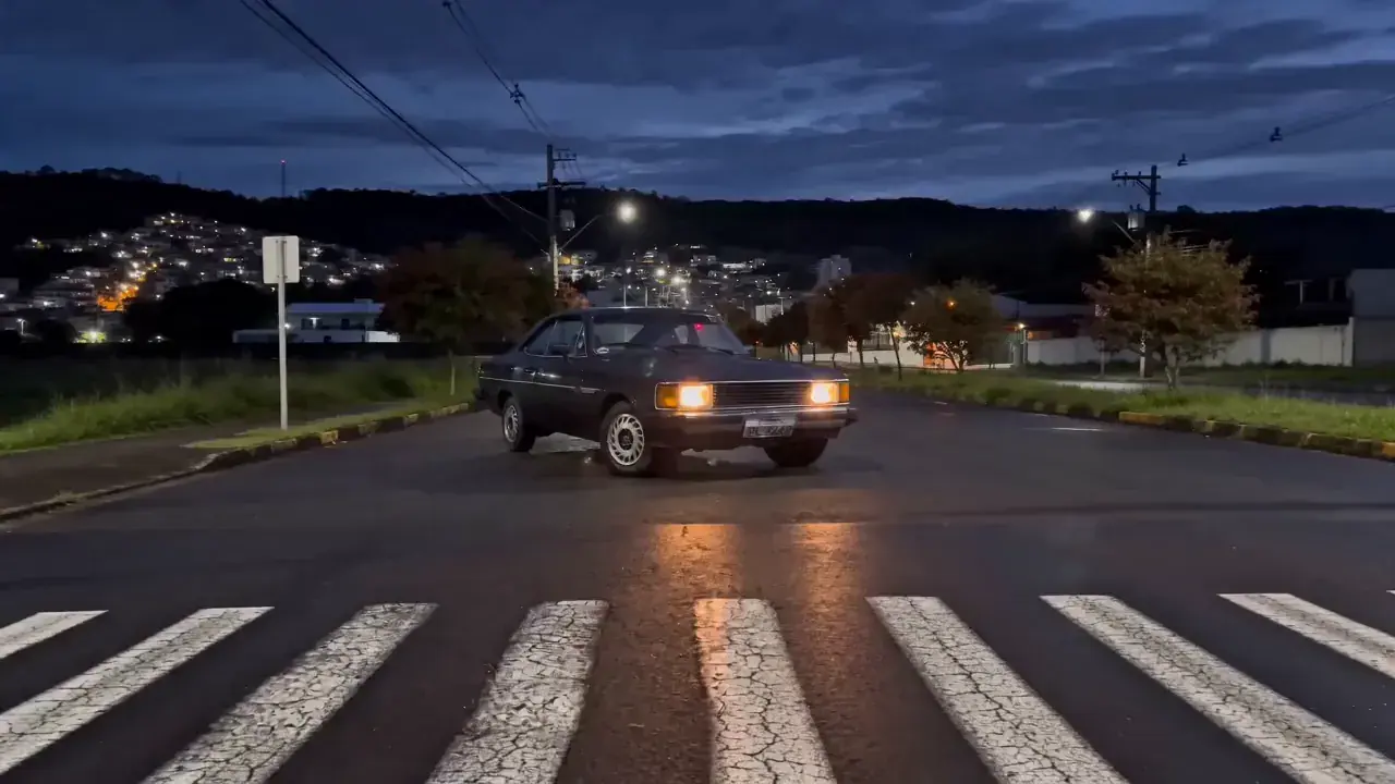 Opala preto atravessando a rua com os faróis acesos ao entardecer.