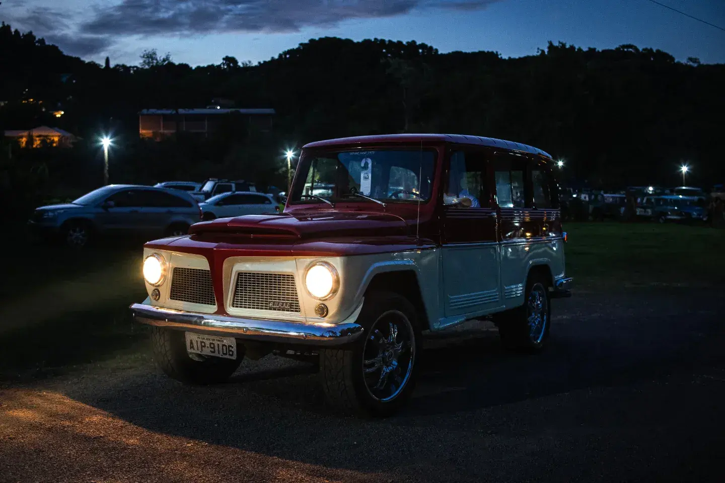 Rural Willys cinza e vermelha com os faróis acesos em um encontro noturno de veículos antigos.